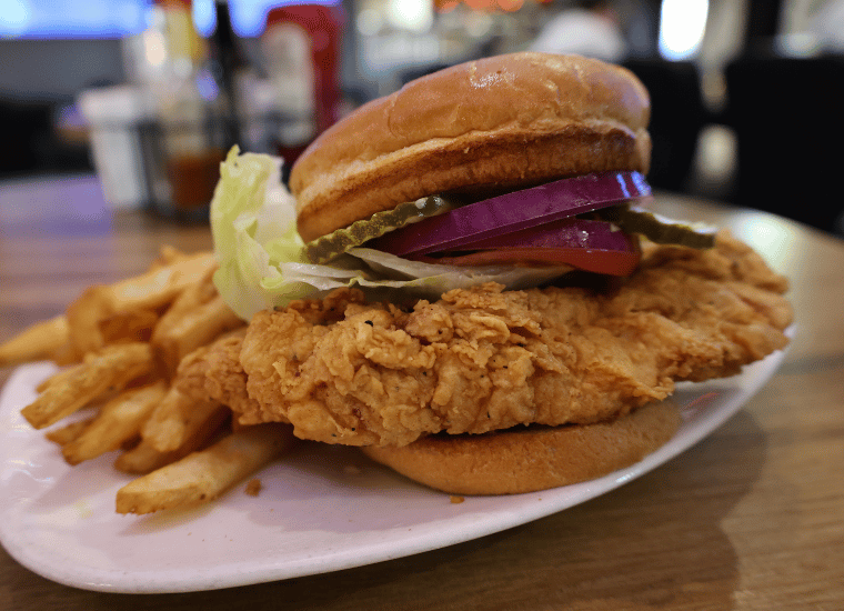 Close-up of Wasted Grain’s award-winning breaded pork tenderloin sandwich on a white plate. The crispy, golden-brown tenderloin hangs past the edges of a toasted bun and is topped with lettuce, tomato, red onion and pickles.