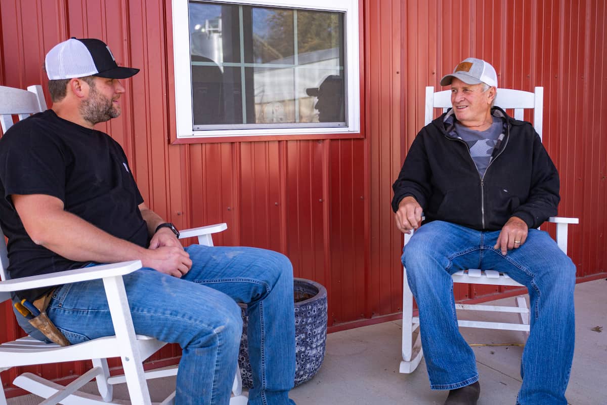 Dad and adult son sit in rocking chairs beside red barn
